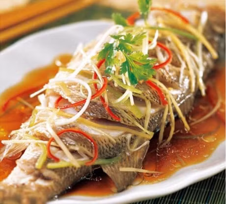 Hòa tan 2 thìa đường vào 2 muỗng canh nước mắm rồi rưới đều lên cá, sau đó cho thêm gừng đã thái sợi vào và hấp trong 5 phút. Cho tỏi băm đã phi thơm, hành lá thái khúc rồi cho vào nồi cá đang hấp. Ảnh: nghethuatamthuc.vn.
