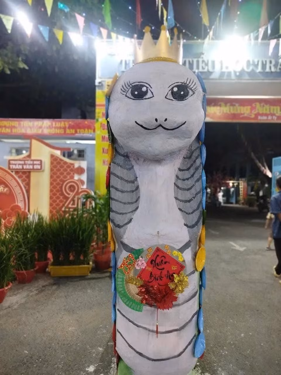 "Bé Na" trước cổng Trường Tiểu học Trần Văn Ơn (Biên Hòa, Đồng Nai) gây cười với vẻ ngoài giống nữ hoàng Ai Cập. Chia sẻ với Tri Thức - Znews ngày 13/1, chị Đào Thị Hằng (thành viên Hội phụ huynh học sinh trường) cho biết đây là tác phẩm hội tặng cơ sở con đang theo học nhằm góp vui cho dịp Tết Nguyên Đán. Tượng được làm từ xốp trắng để tạo hình, bên ngoài là đĩa ghép lại thành vảy rắn. Để trông đỡ đơn địu, chị Hằng trang trí thêm vương miện, vẽ mi cong, mắt long lanh và treo hoa với dòng chữ "Xuân bình an" trước ngực. Ảnh: Trường Tiểu học Trần Văn Ơn.