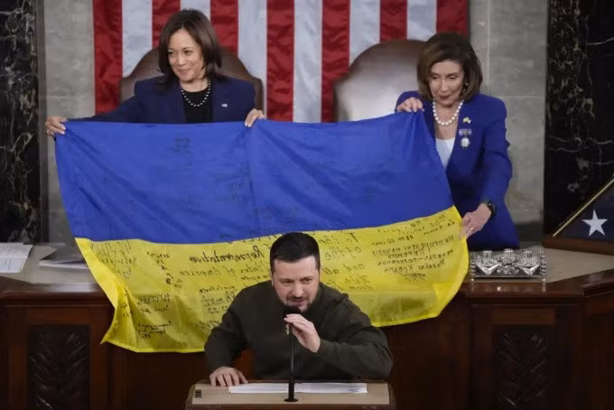 Tổng thống Ukraine Volodymyr Zelensky phát biểu trong khi Phó tổng thống Mỹ Kamala Harris (trái) và Chủ tịch Hạ viện Nancy Pelosi giăng lá cờ Ukraine phía sau tại thủ đô Washington, Mỹ ngày 21/12. Ảnh: AP.