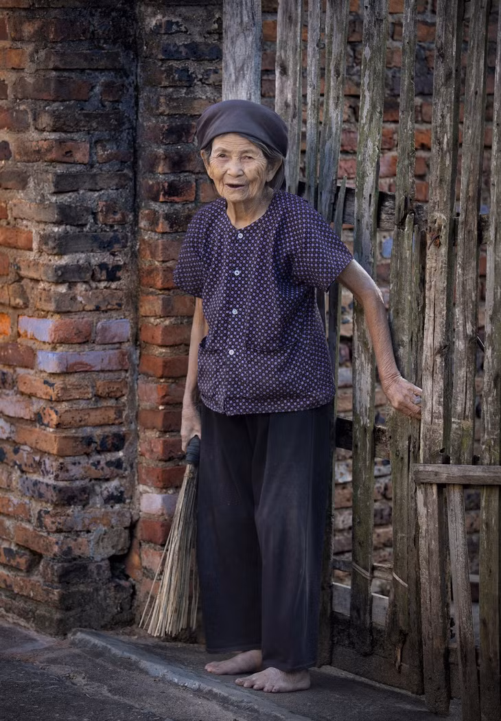 Cụ bà bên bức tường nhuốm màu thời gian, nở nụ cười, khoe hàm răng đen khi gặp ống kính. Phong tục nhuộm răng đen có từ xưa, ngày càng ít người giữ được truyền thống này.